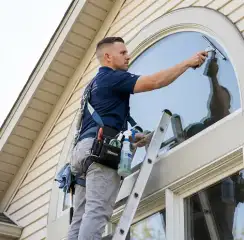 Professional window cleaner on a ladder using a squeegee to clean an exterior arched window, Kansas City, MO.